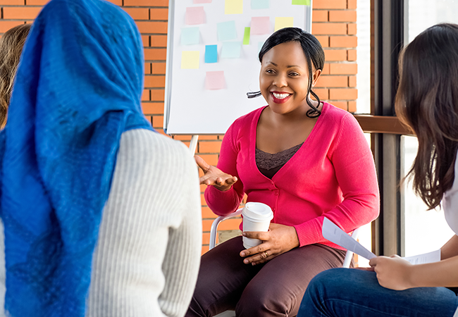 A young African-American woman sits among two other women, one wearing a hijab, and leads a meeting in front of a whiteboard.